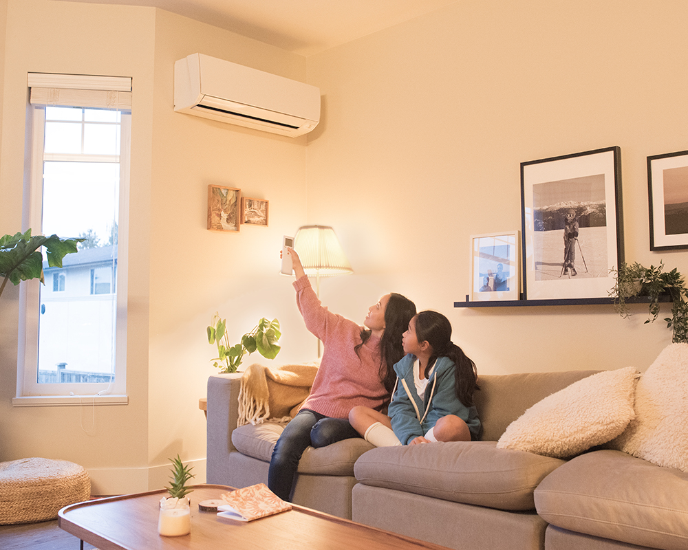 A mother and daughter are sitting on their living room couch. The mother is pointing a remote at the heat pump installed on the wall behind her.